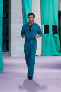 Model wearing leather bomber jacket in teal, with a piqué polo shirt in black and leather trousers in teal.