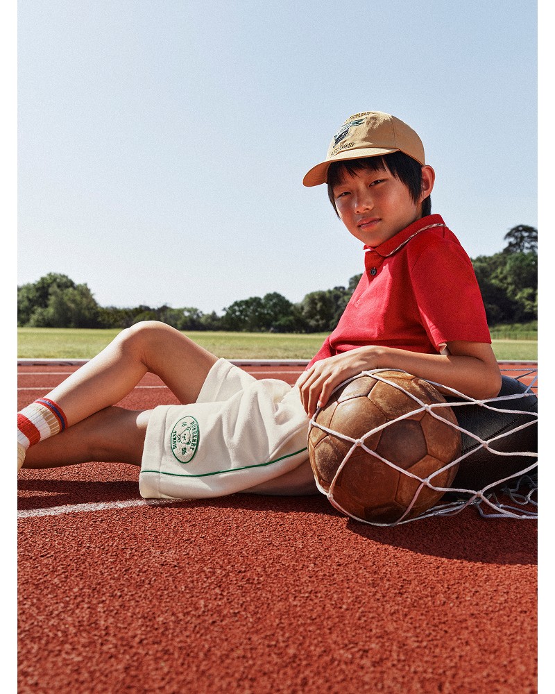模特身穿亮红色男童Polo衫，佩戴沙色学院风棒球帽